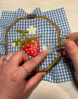 Felting embroidery hoop with blue gingham fabric, strawberry motif, needle being worked by hands