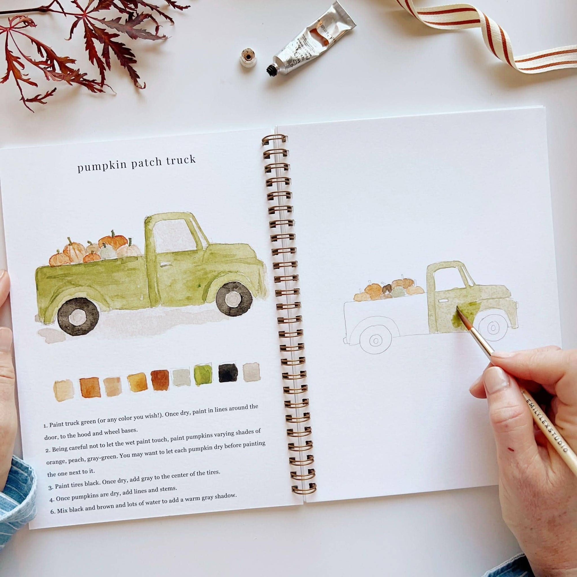 Autumn watercolor workbook open to pumpkin patch truck painting page with color swatches