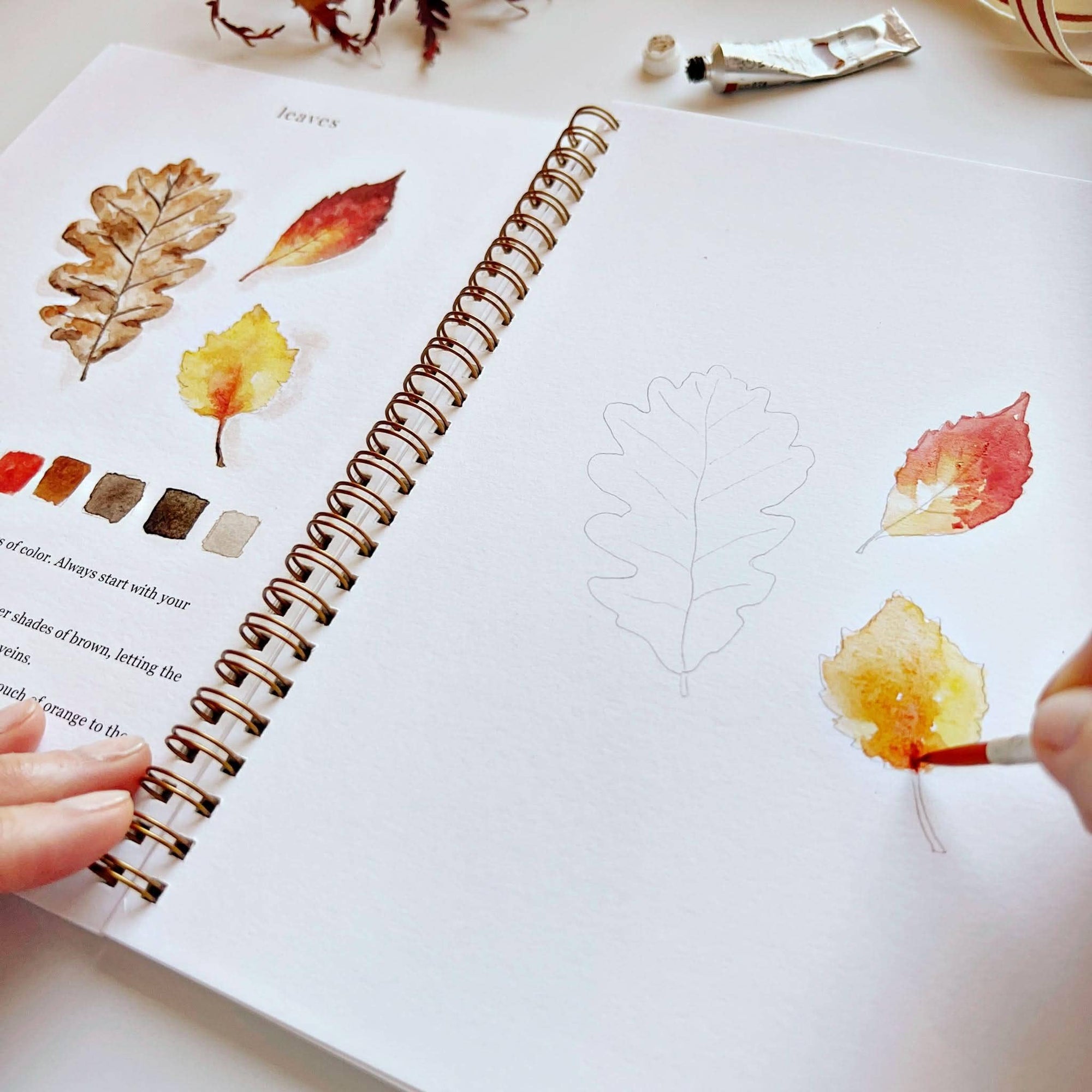 Autumn watercolor workbook open page with leaf paintings and brush applying watercolor