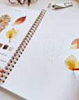 Autumn watercolor workbook open page with leaf paintings and brush applying watercolor